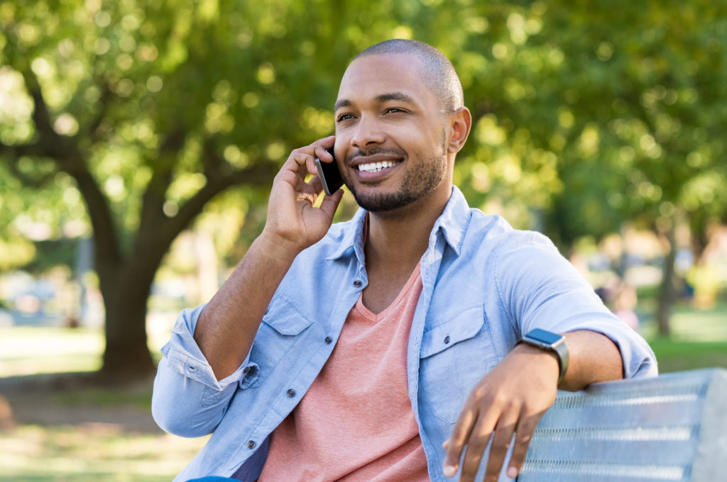 Handsome african man talking on phone at park. Young black man talking on cellphone while sitting on bench outdoor. African american smiling guy in a happy conversation at mobile phone.