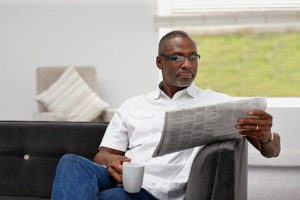 Smart afro american mature man wearing smart glasses sitting on sofa reading newspaper with a cup of coffee